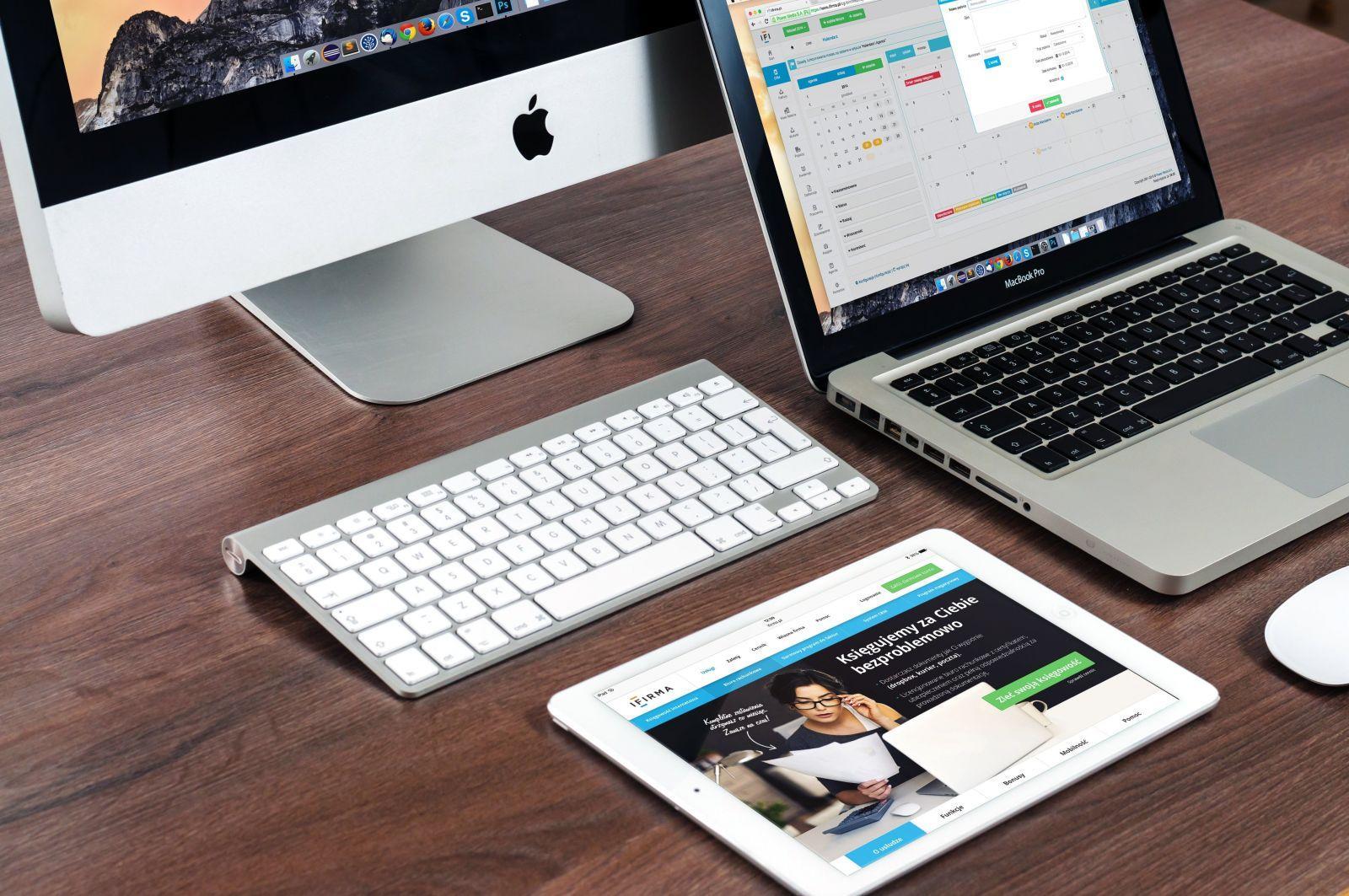 Workspace with Apple iMac, MacBook Pro, white wireless keyboard and a tablet displaying a business website on a wooden desk