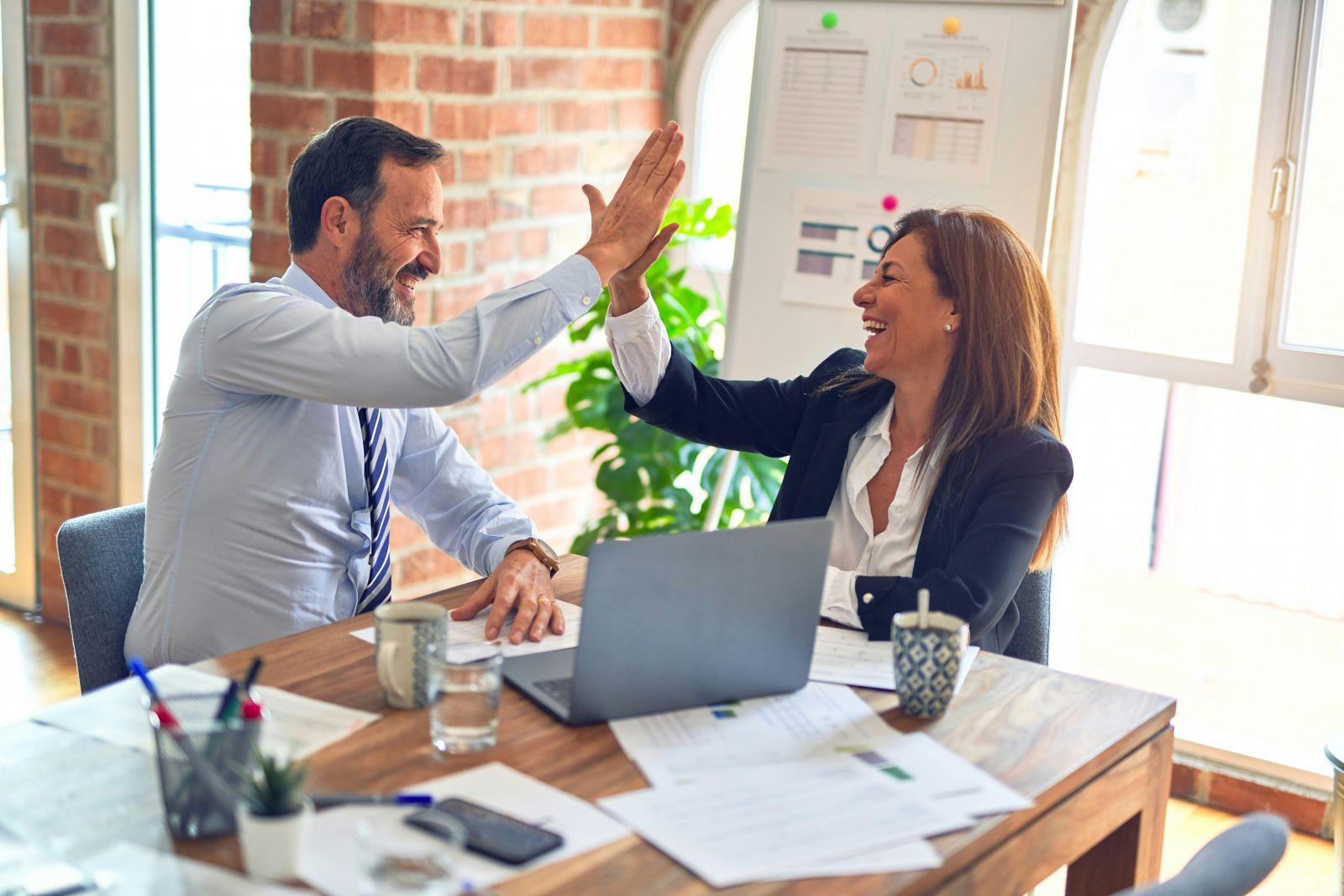 Two business colleagues smiling and high-fiving each other at a meeting table with laptop and documents