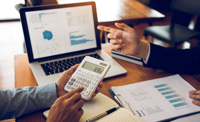 Two people discussing financial charts shown on a laptop screen and printed reports with one using a calculator