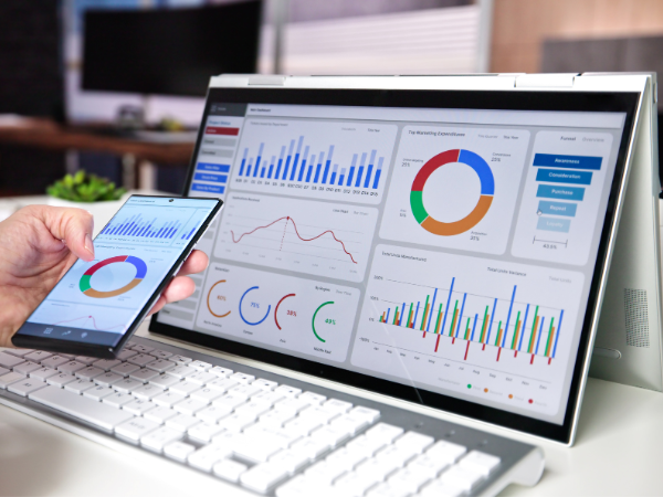 Hand holding smartphone displaying data dashboard matching charts on computer monitor, showing bar charts, pie chart and line graphs in a workspace