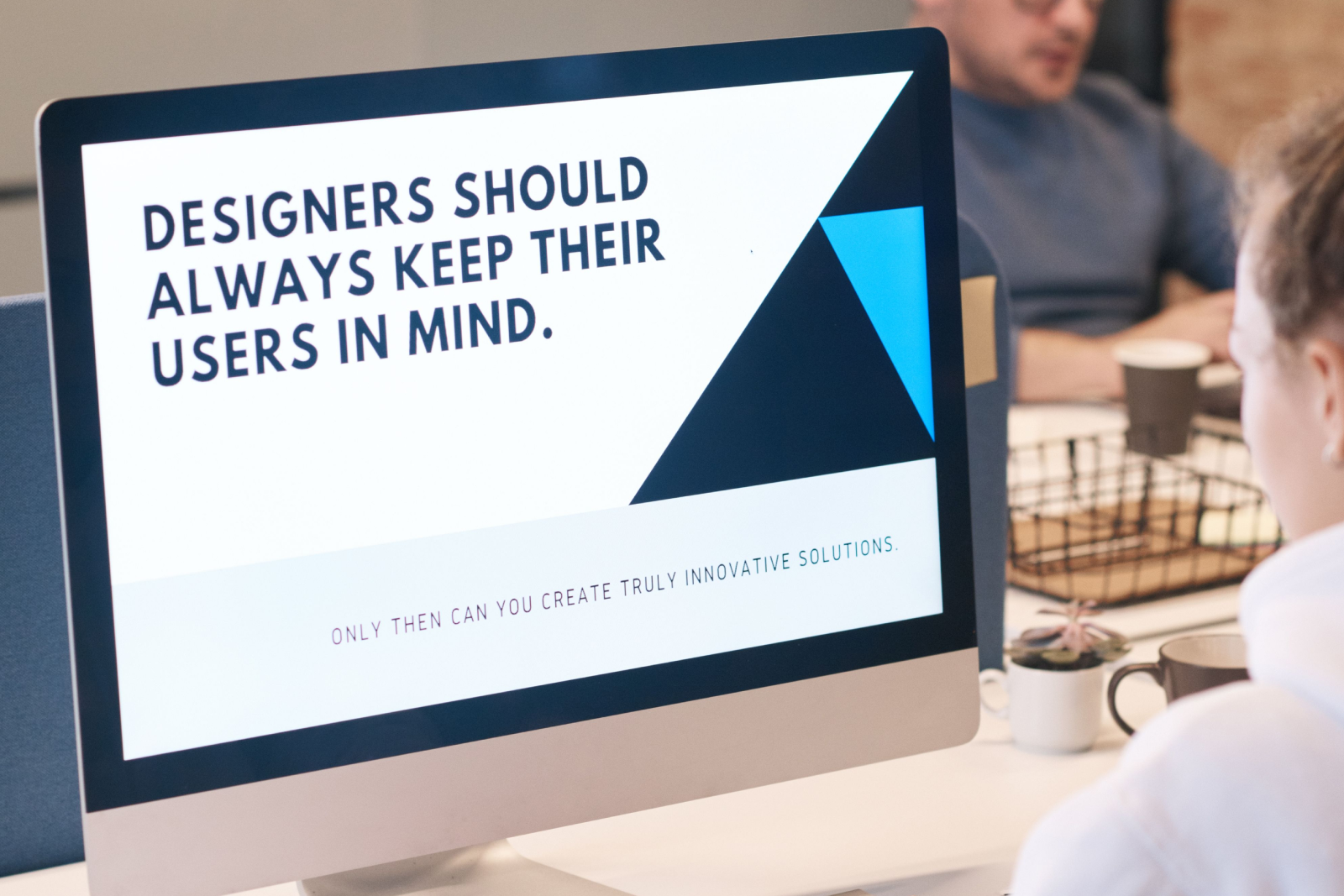Computer screen displaying the phrase 'Designers should always keep their users in mind' with geometric shapes, viewed in an office setting
