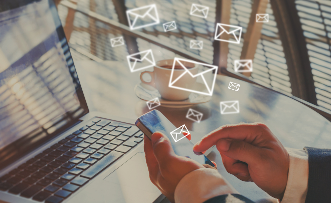 Hand using smartphone with digital email icons above near laptop and coffee cup on table