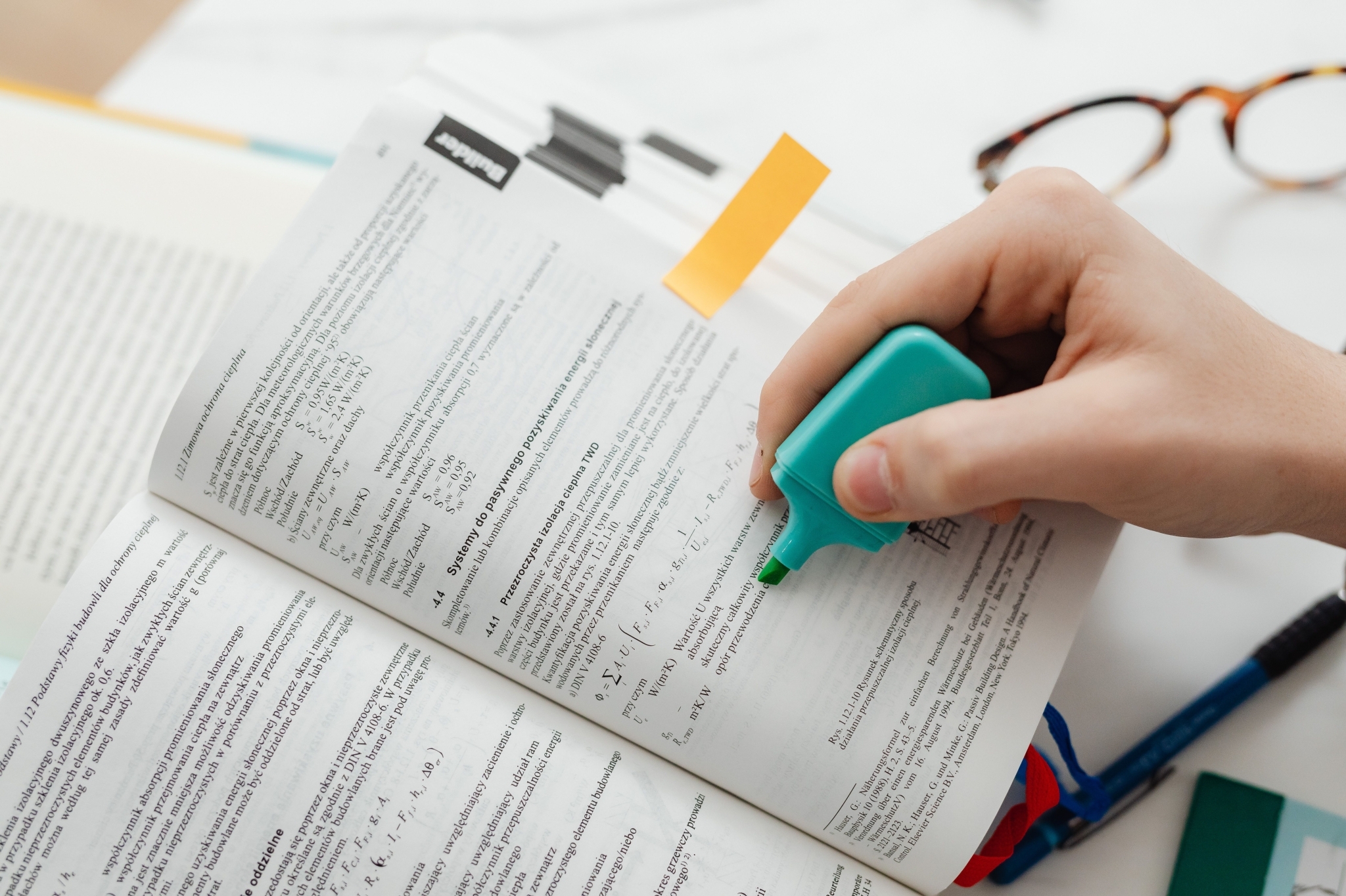 Person holding a turquoise highlighter and marking text in an open book with glasses and pens nearby