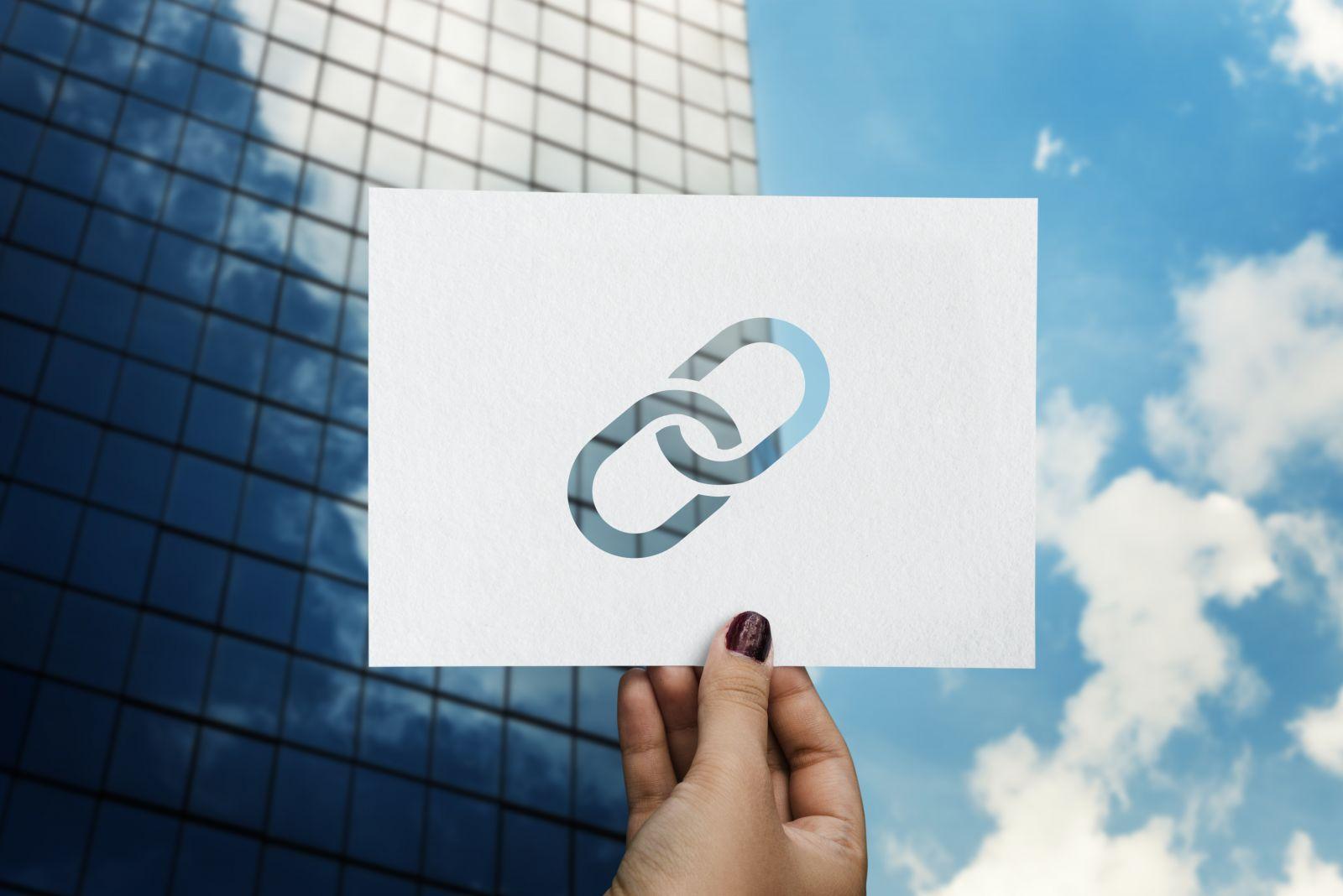 Hand holding white paper with a cutout link icon showing blue sky and glass building behind