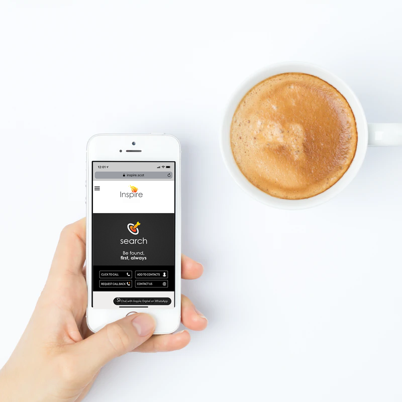 Hand holding a white smartphone displaying the Inspire website search page next to a cup of coffee on a white surface