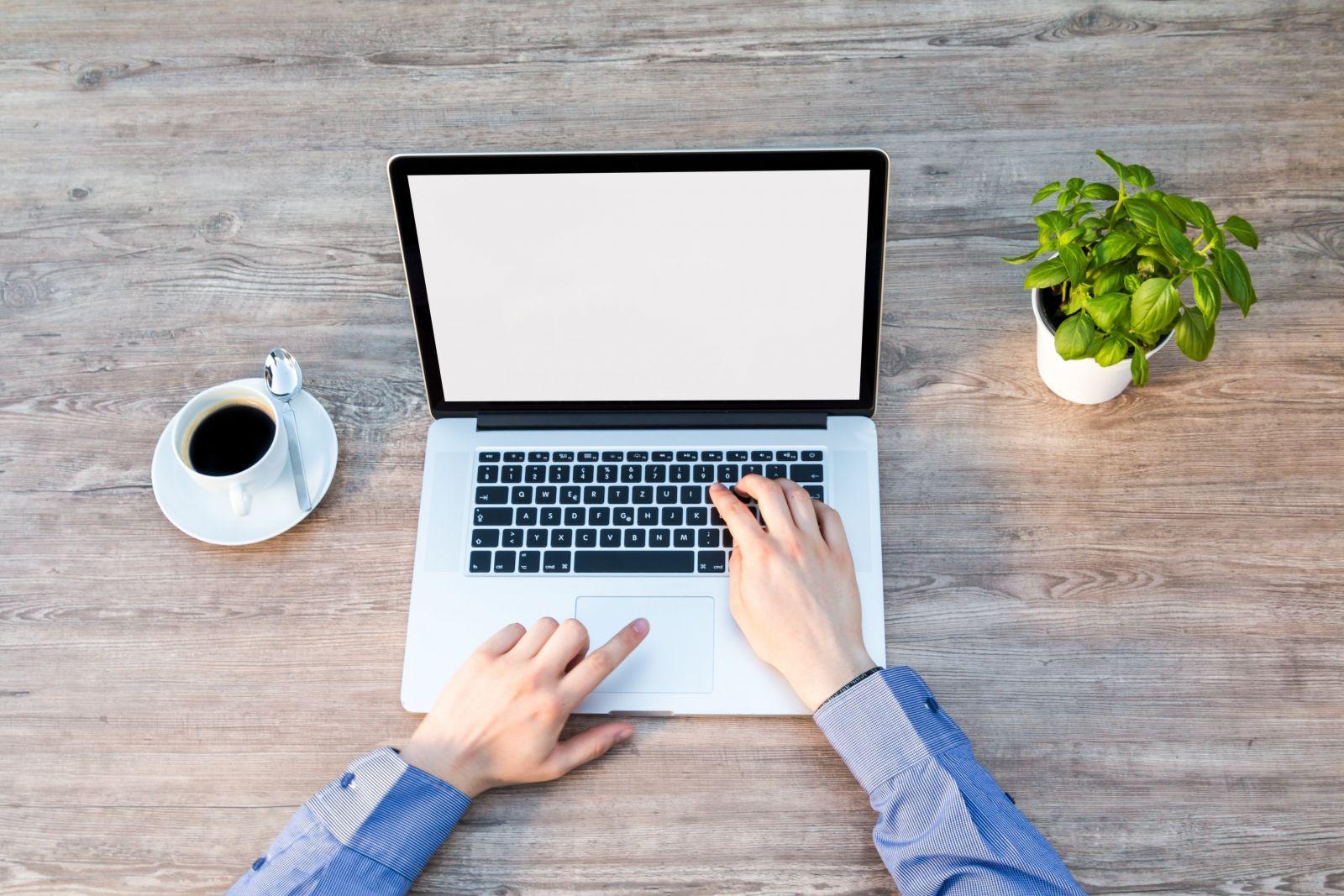 Hands typing on a laptop keyboard on a wooden table next to a cup of coffee and a potted green plant