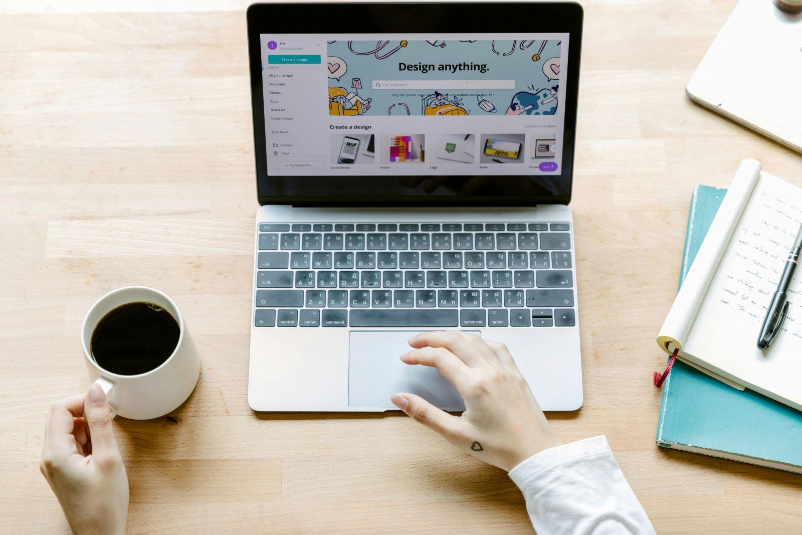 Person using a laptop with a design application open on screen, holding a cup of coffee, with notebooks and a pen on the wooden desk