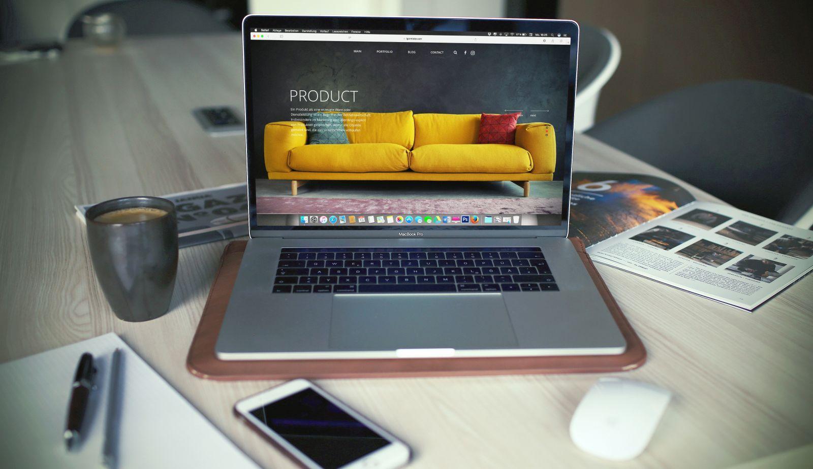 Open laptop on a desk displaying a website with a yellow sofa product image, alongside a coffee cup, smartphone, pen, and magazine