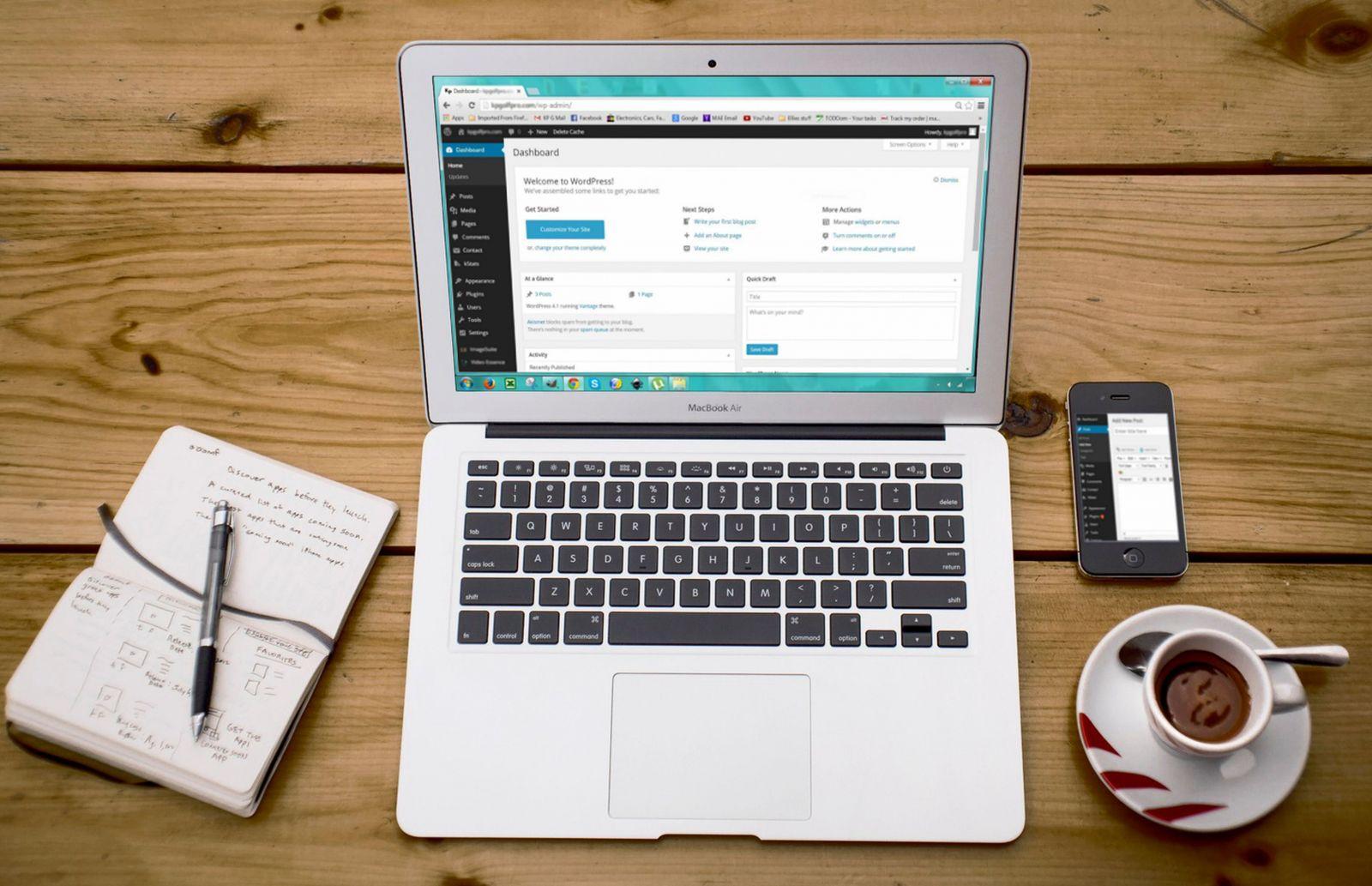MacBook Air laptop displaying WordPress dashboard on screen, beside a notepad with pen, smartphone showing blog post interface, and cup of espresso on wooden table