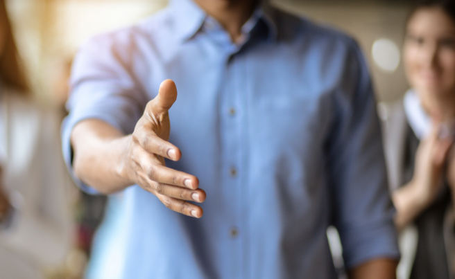Man wearing a blue shirt extending his hand for a handshake with blurred people in the background