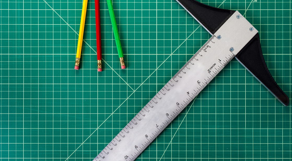 Metal T-square ruler and three coloured pencils with erasers on a green cutting mat with grid lines