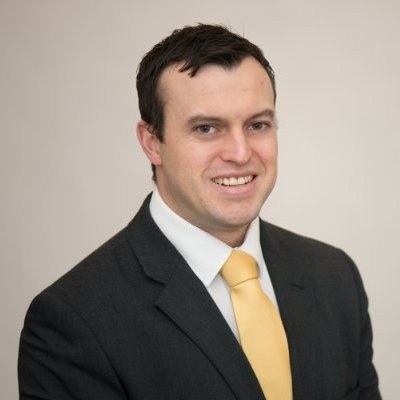 Portrait of a man in a dark suit and yellow tie smiling against a plain background