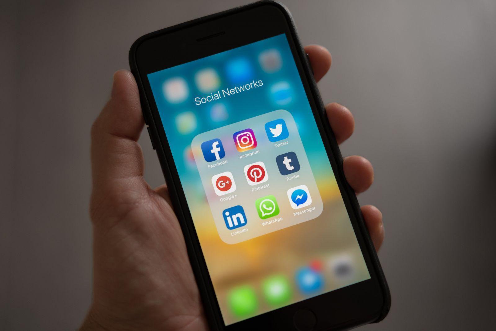 Hand holding a smartphone displaying a folder named Social Networks with apps including Facebook, Instagram, Twitter, Google+, Pinterest, Tumblr, LinkedIn, WhatsApp, and Messenger