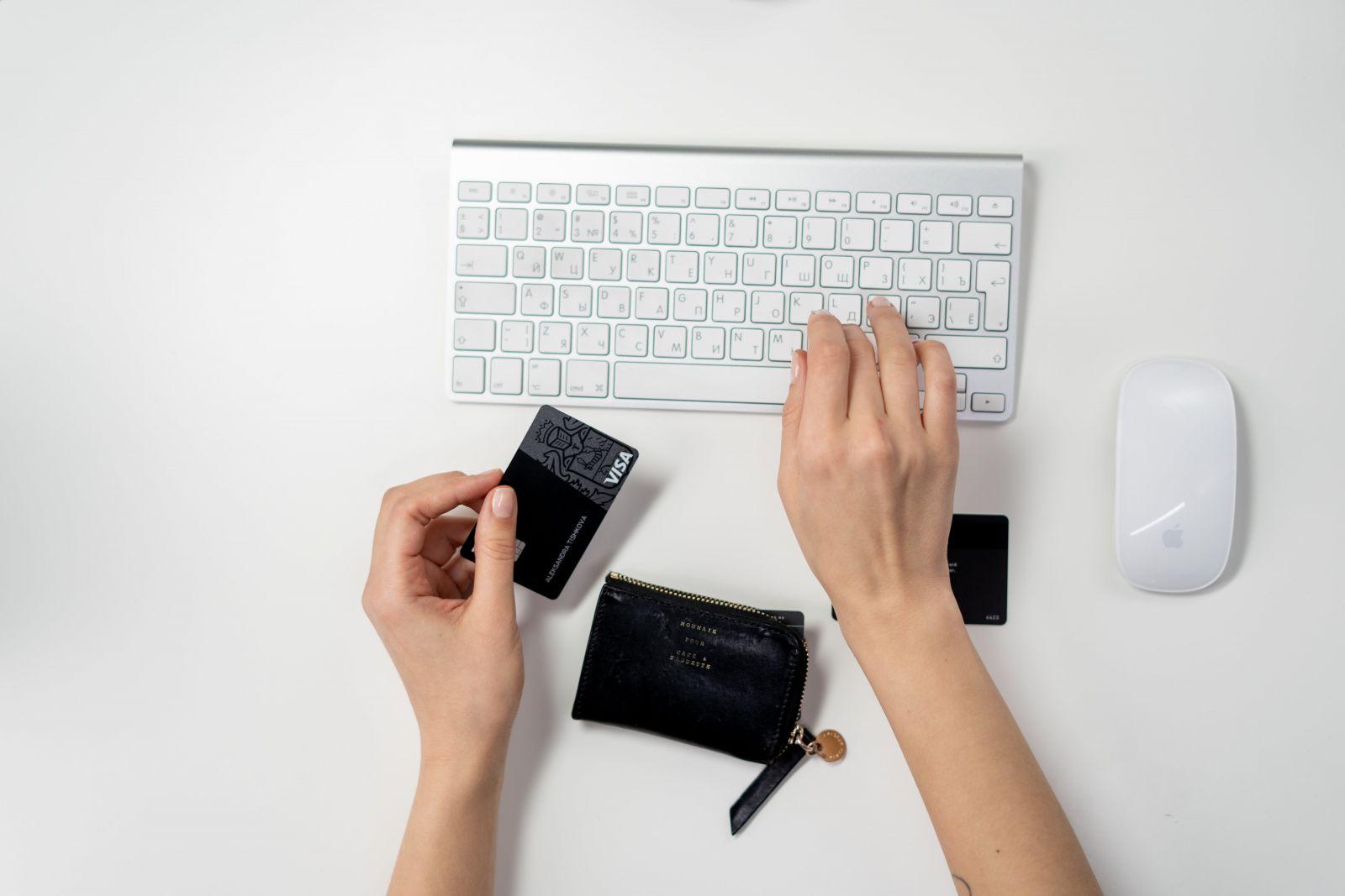 Hands holding a black Visa credit card and typing on a white keyboard next to a black wallet and white Apple mouse on a white desk