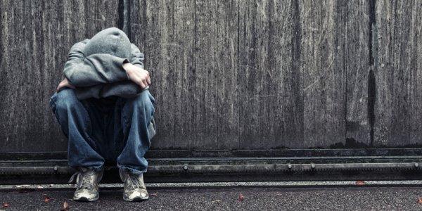 Person sitting on the ground with head down and covered by hoodie, against a rough wall outdoors