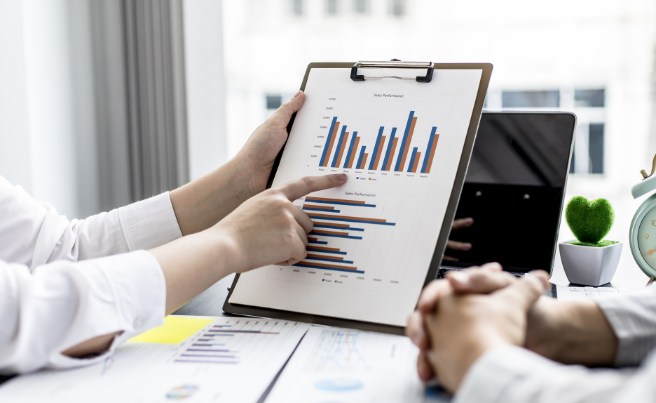 Person pointing at sales performance bar charts on a clipboard during a business discussion