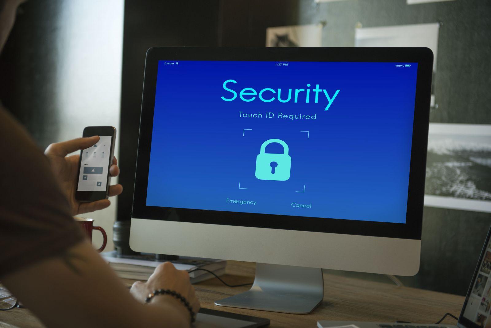 Computer screen displaying a security lock screen requiring Touch ID verification with a padlock icon, person holding a smartphone nearby