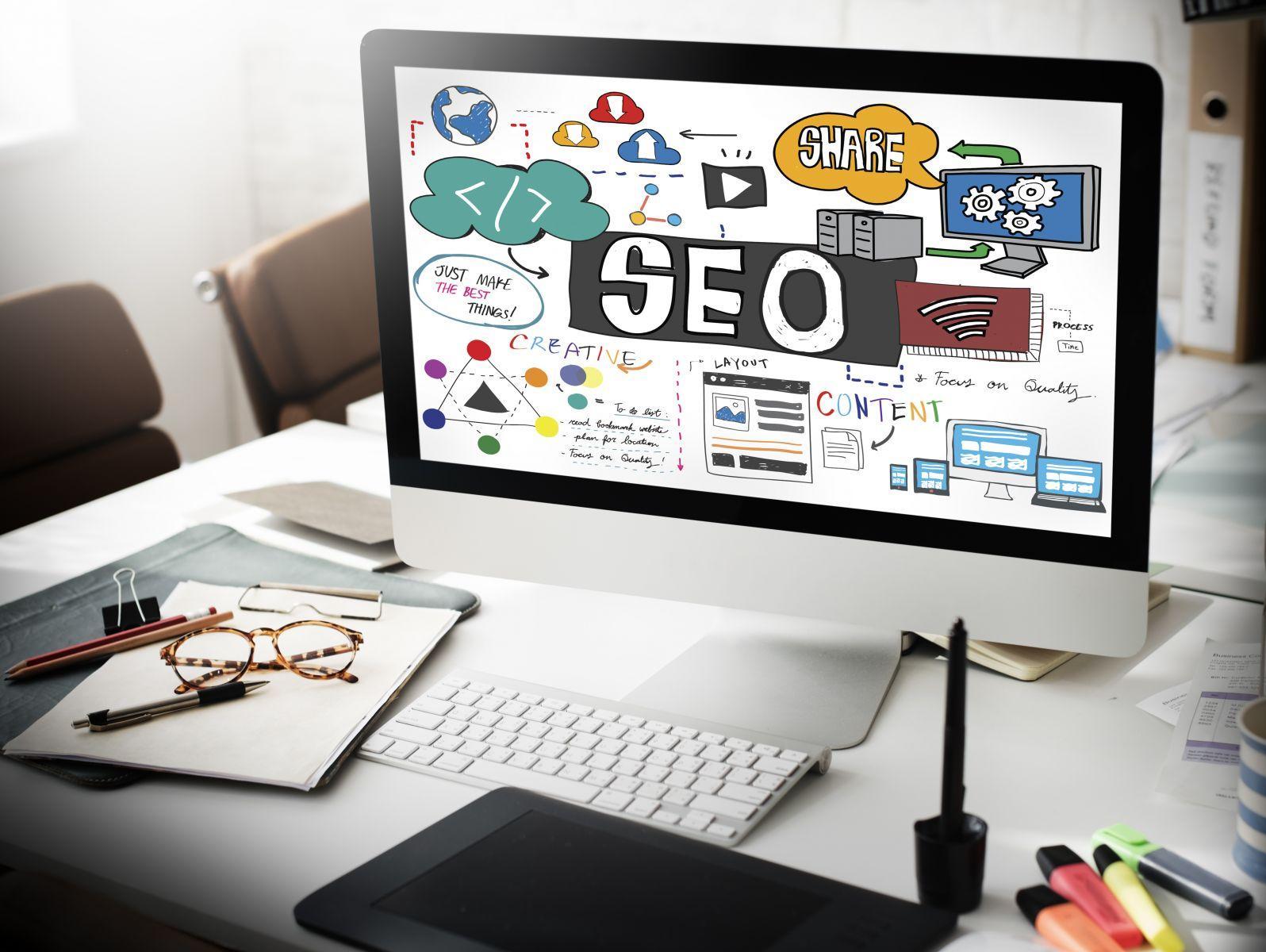 Computer screen showing colourful sketches and text illustrating SEO concepts including content, share, creative, and layout in a workspace with office supplies