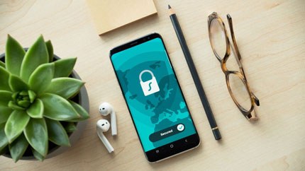 Smartphone displaying a password screen with a lock icon on a wooden desk alongside a pencil, glasses, wireless earphones, and a small succulent plant