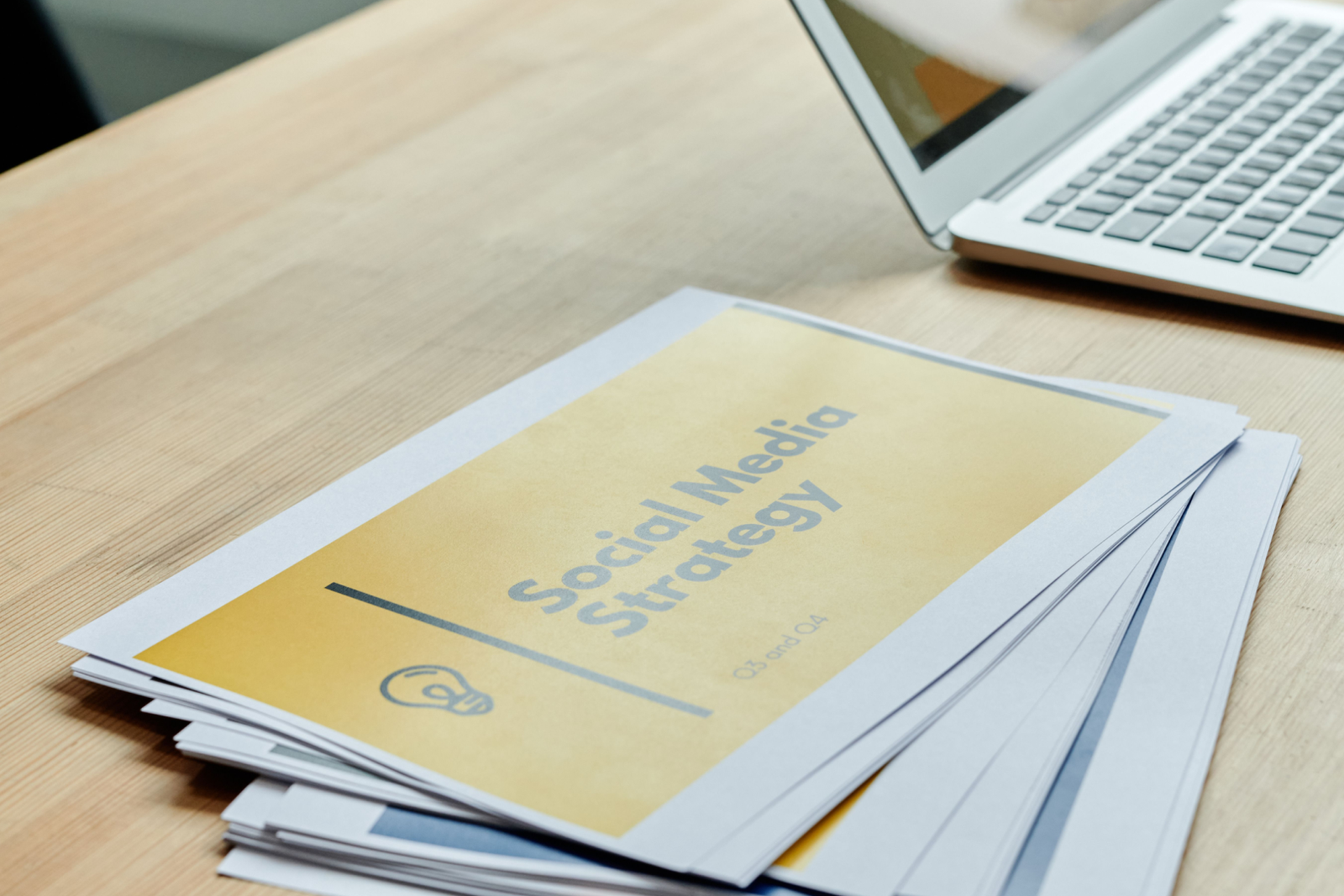 Stack of printed documents titled Social Media Strategy on a wooden desk beside a laptop