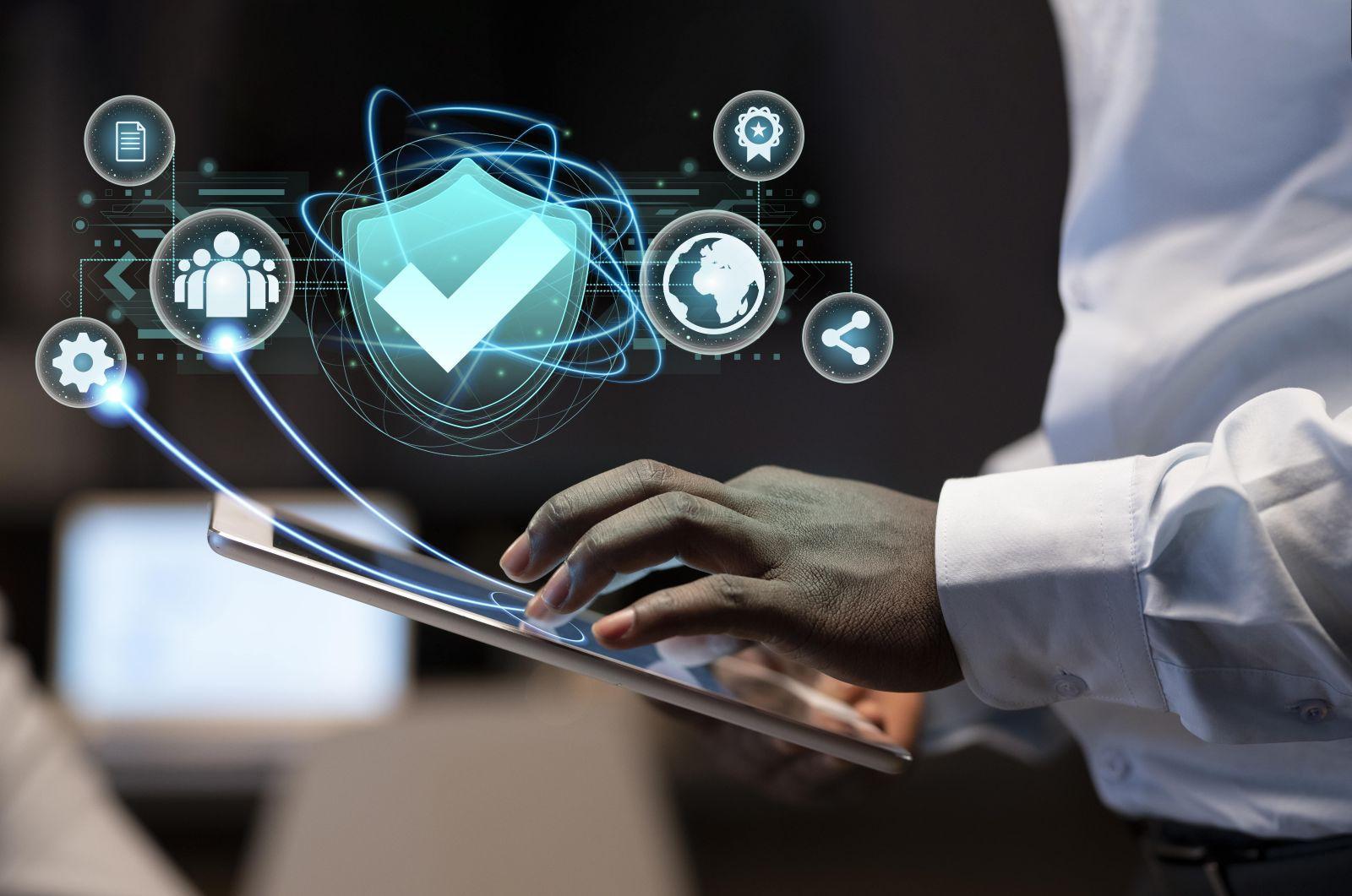 Hand using a tablet with digital security icons including a shield with a checkmark, people, a globe, and sharing symbols floating above it