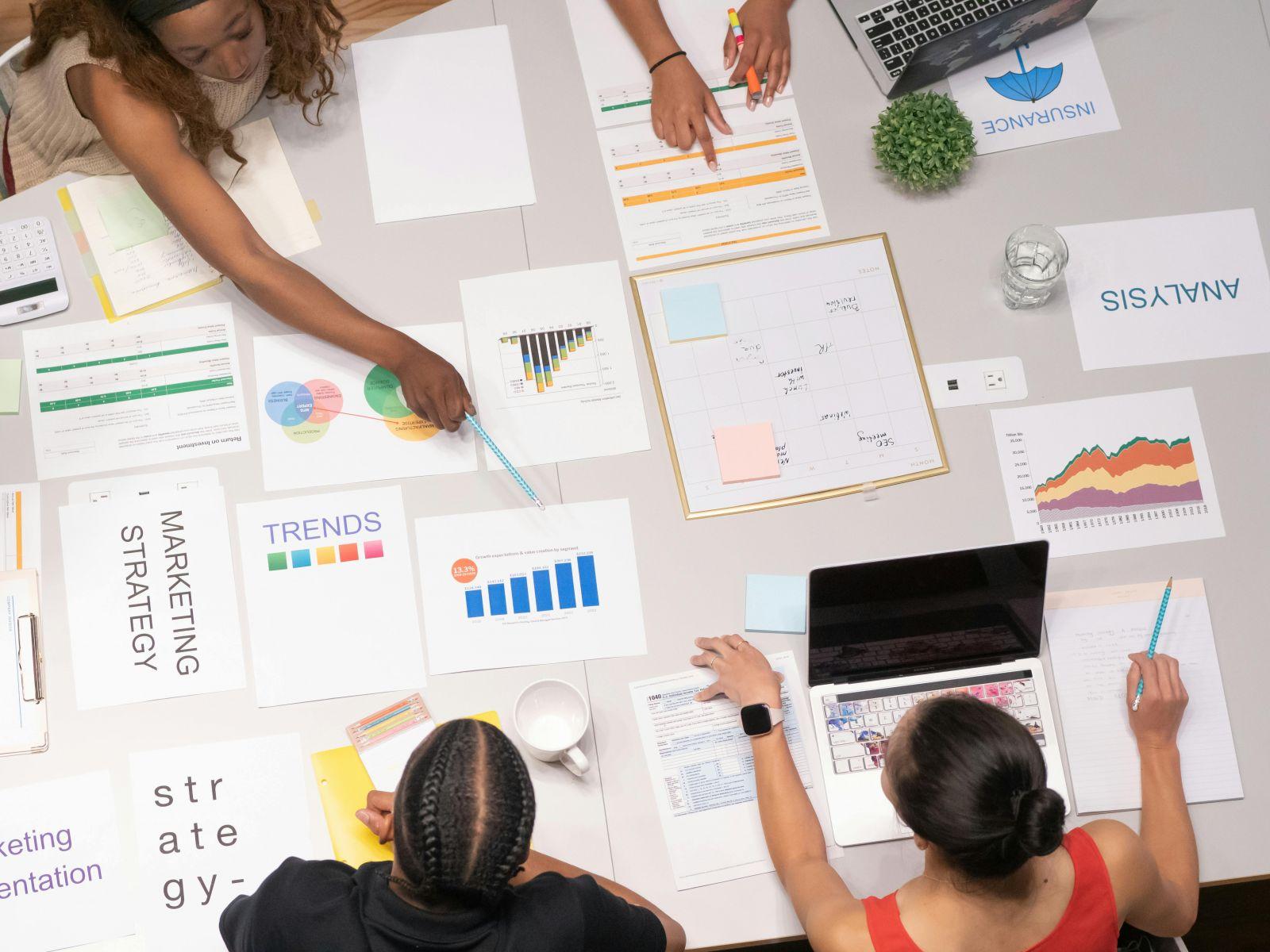 Overhead view of three people collaborating on marketing strategy with charts, graphs and notes spread on a table