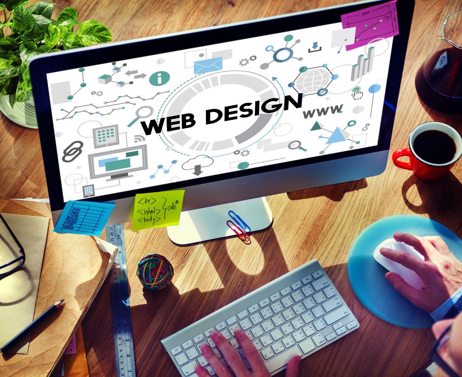 Desktop computer screen displaying web design graphics and text, with hands using keyboard and mouse on a wooden desk with stationery and a coffee cup
