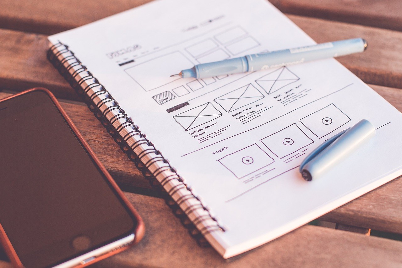 Notebook with website wireframe sketches drawn in pen, accompanied by a smartphone on a wooden table