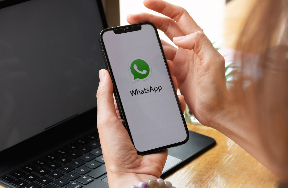 Person holding a smartphone displaying the WhatsApp logo on screen with a laptop in the background