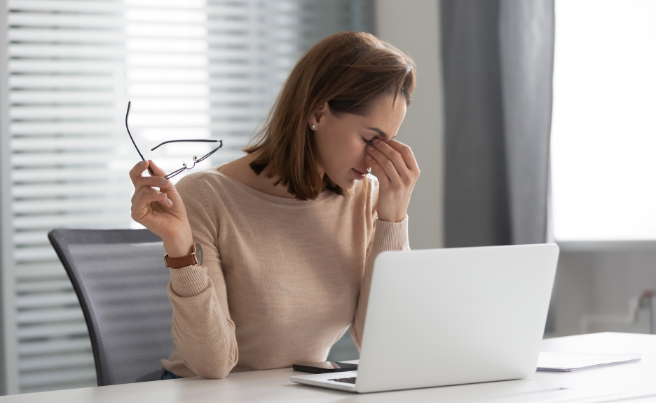 Woman sitting at desk with laptop, holding glasses and rubbing eyes, appearing fatigued or stressed