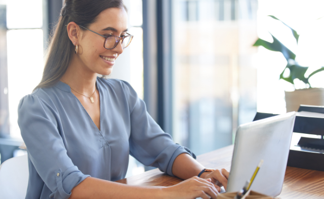 Woman wearing glasses typing on a laptop at a desk in a bright office space