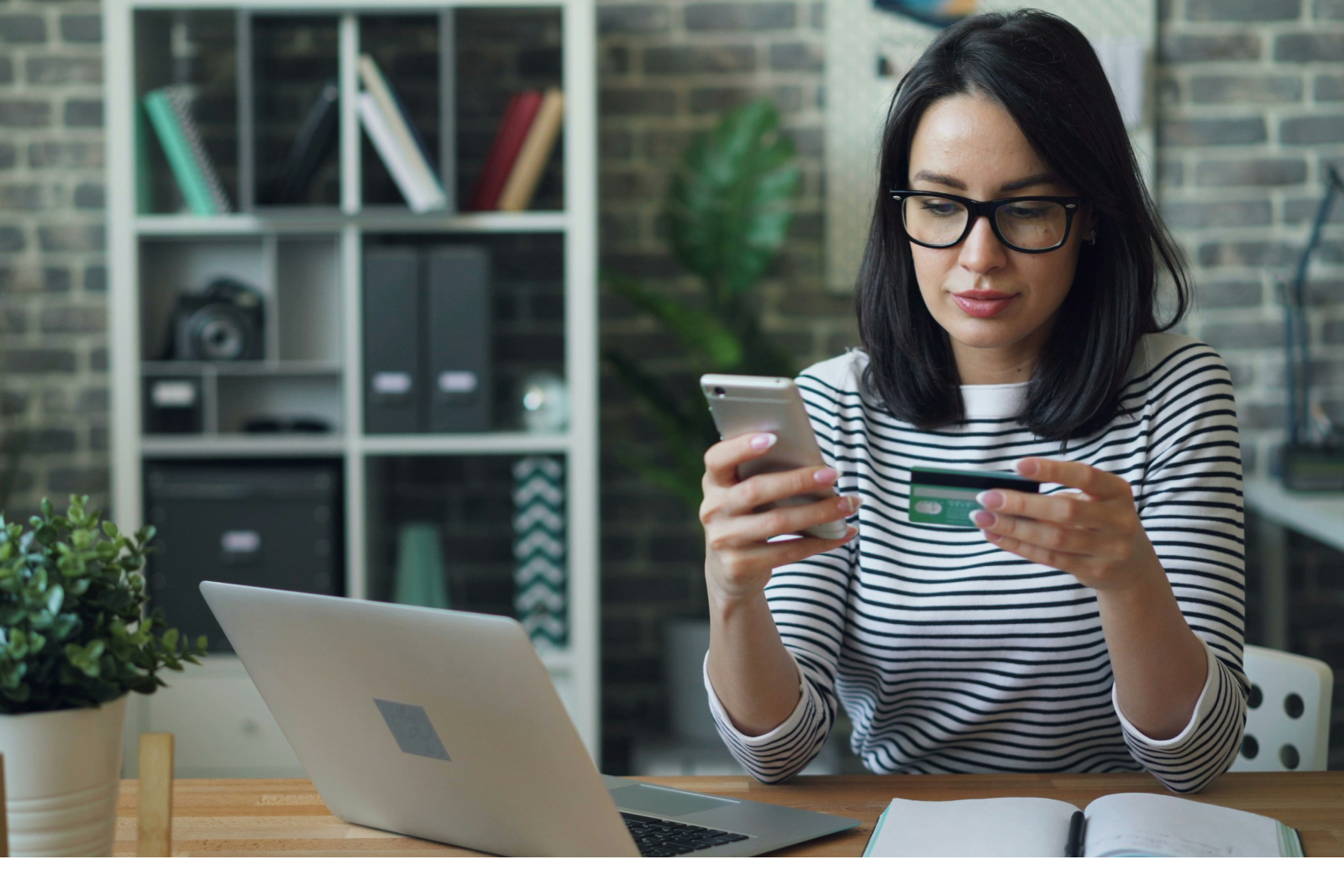 Woman holding a credit card and using a smartphone for an online transaction at a desk with a laptop and notebook