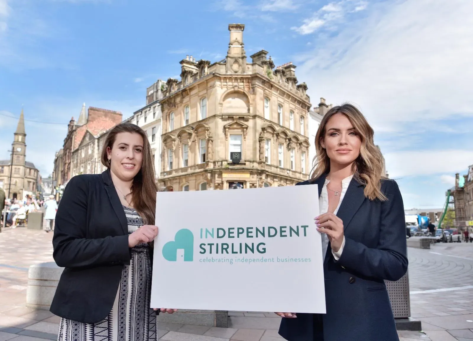 Two women holding a white board displaying the Independent Stirling logo with a historic building in the background