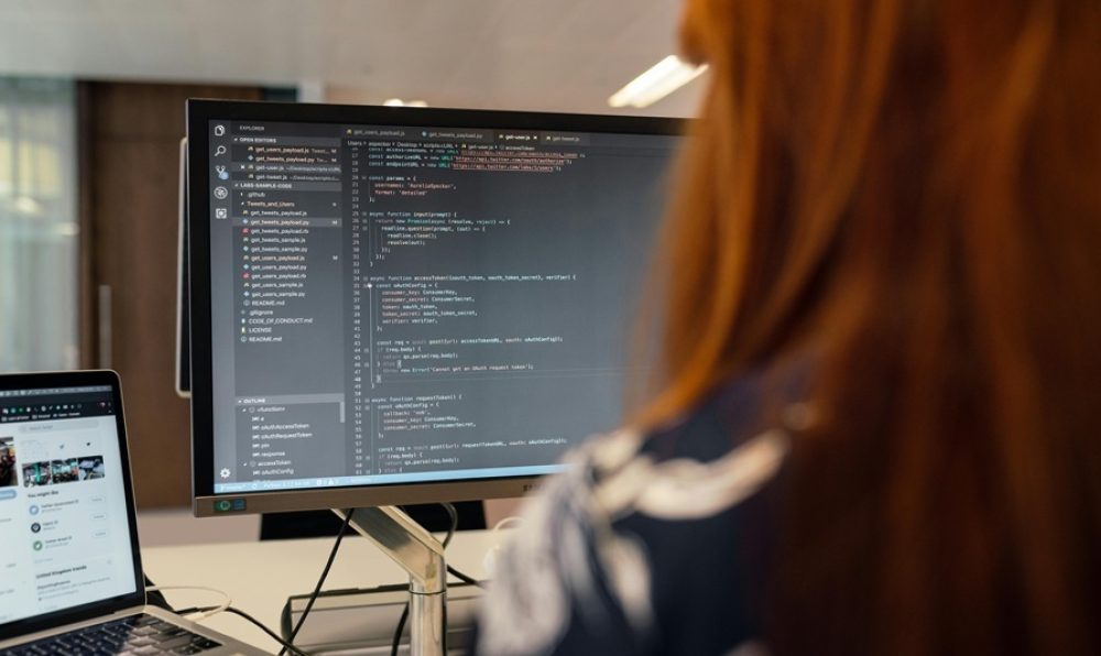Developer working at a desk with code displayed on a large monitor and laptop in a software development environment