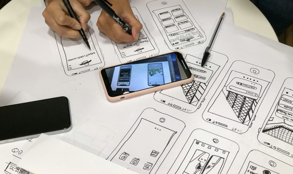 Hands sketching mobile app wireframe designs on paper with a smartphone displaying digital prototypes.