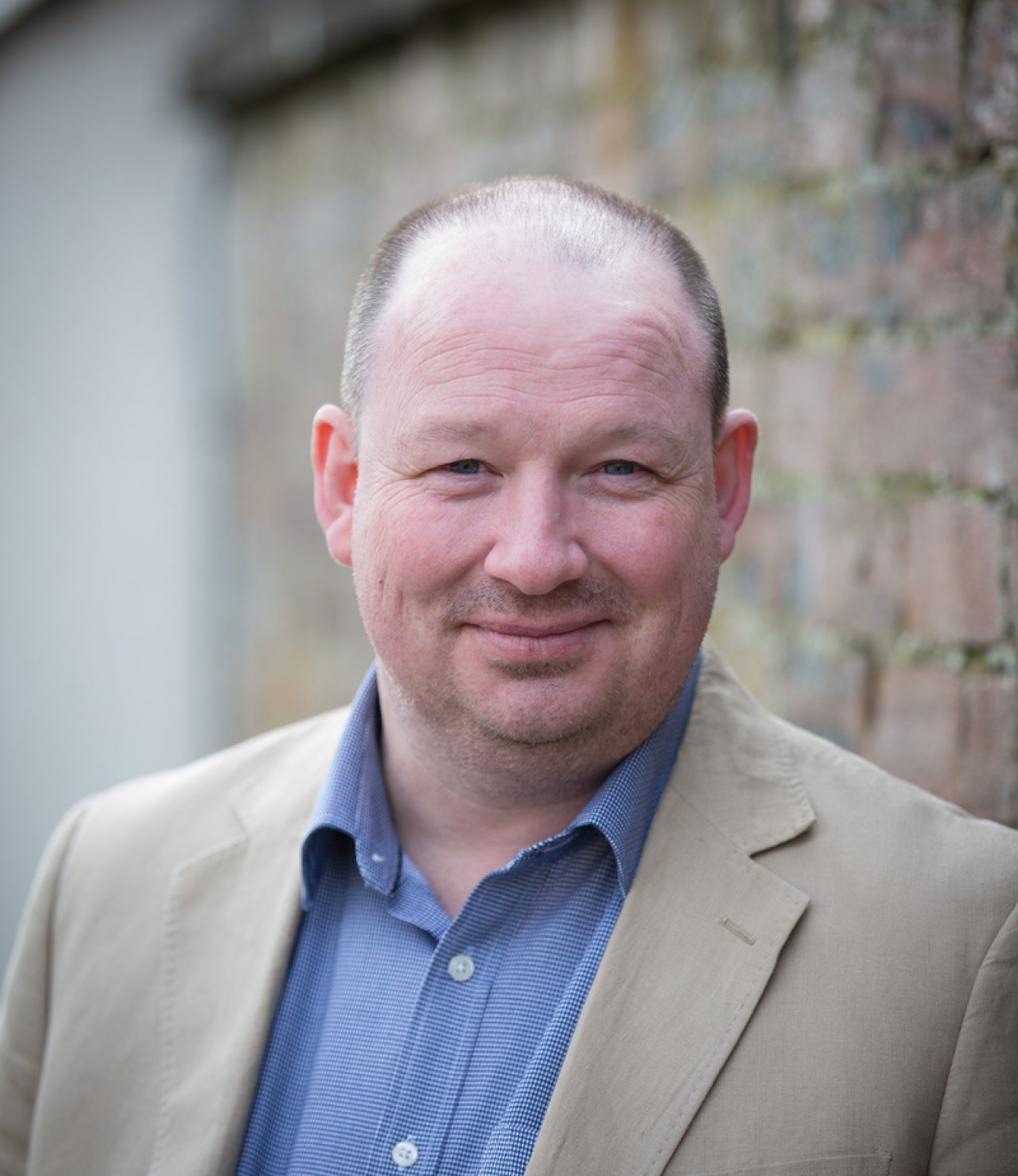 Outdoor portrait of David Dwyer, Expert Witness Lead at Inspire, wearing beige blazer and blue shirt, smiling against a brick wall background.