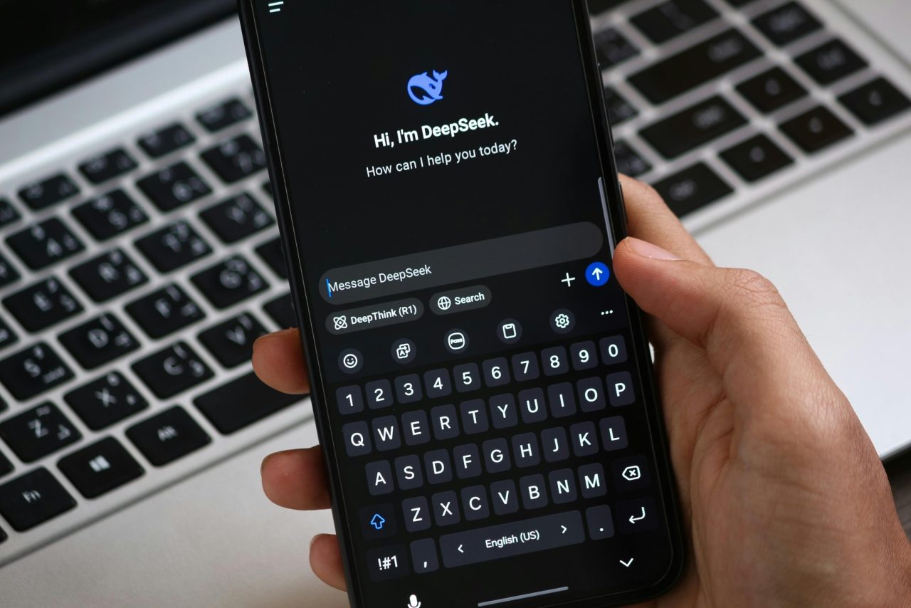 Hand holding smartphone showing DeepSeek AI chat interface with message input and keyboard, laptop keyboard in background