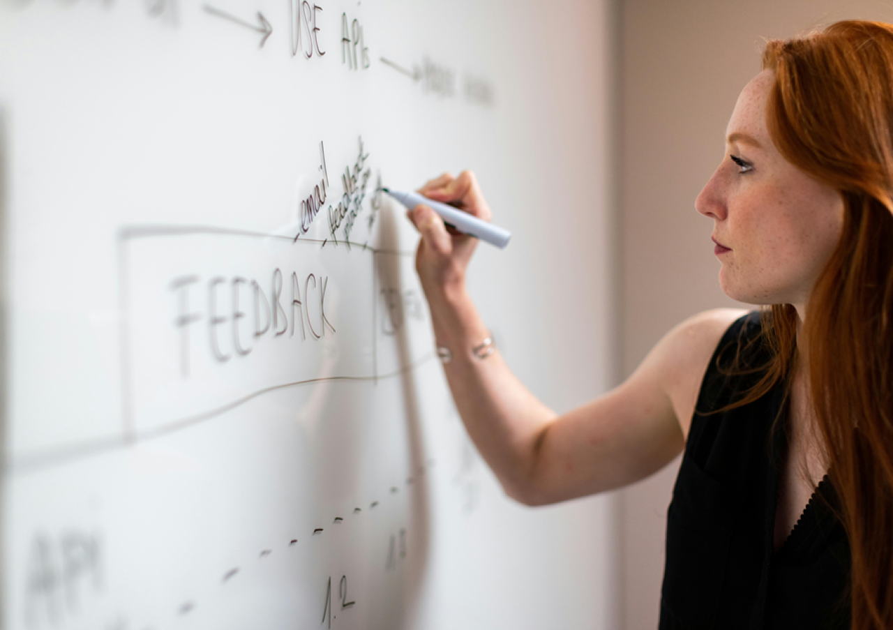 Woman writing a flow diagram on a whiteboard that includes the word feedback and mentions APIs