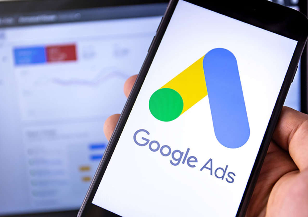 Hand holding a mobile phone showing the Google Ads logo with blurred analytics graphs in the background