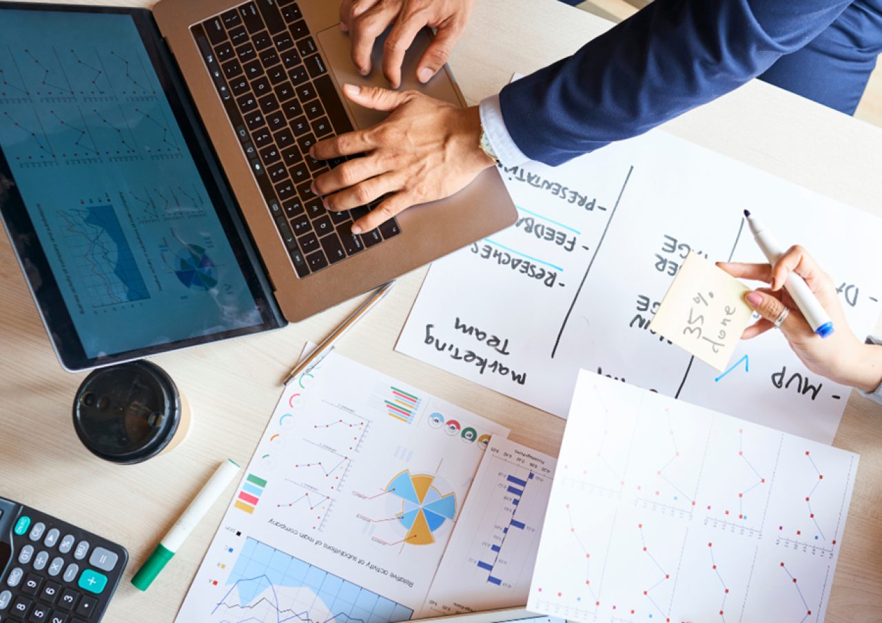 Overhead view of a desk with hands on a laptop and a whiteboard, surrounded by charts and notes related to marketing and paid social campaigns.