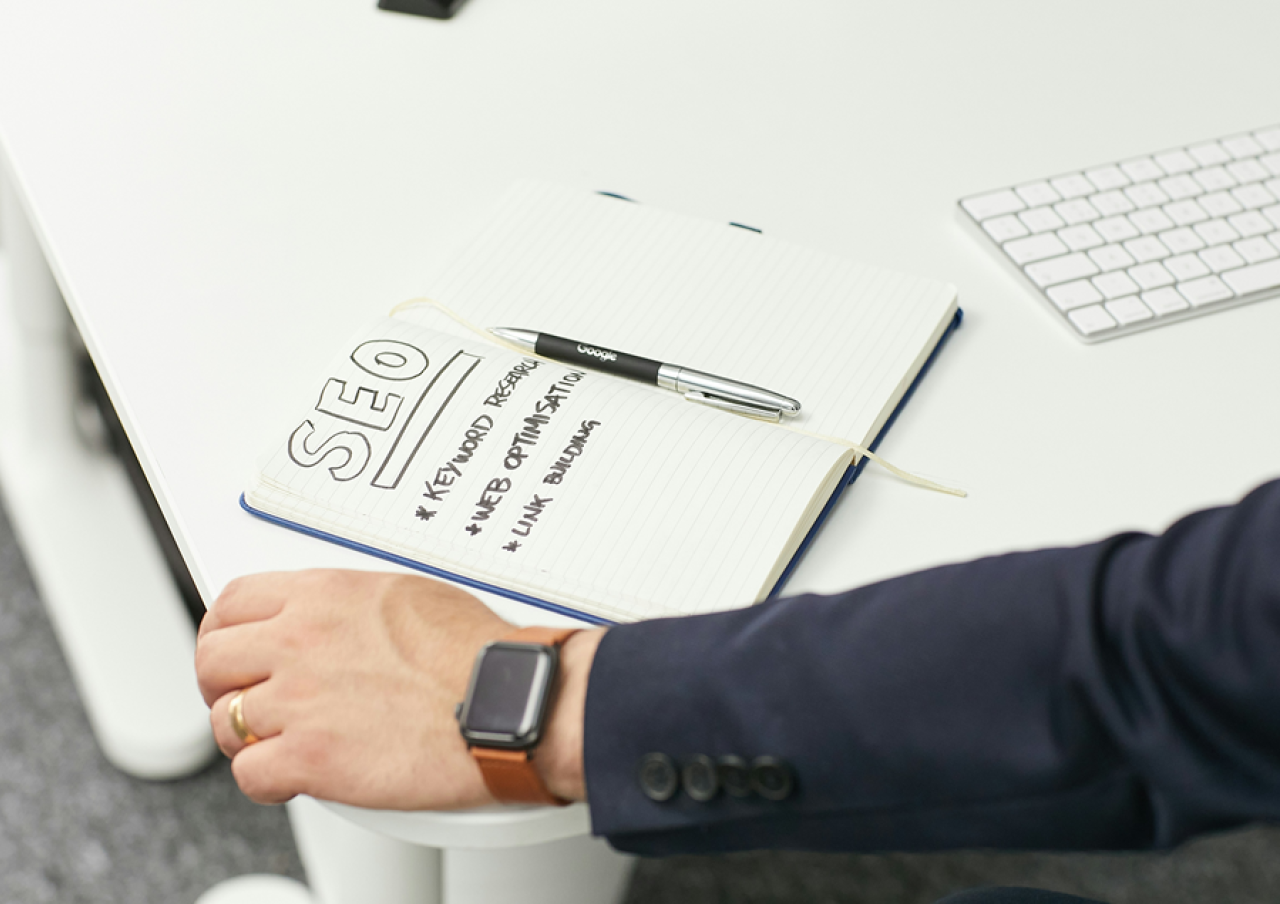 Person's hand near a notepad on a desk with SEO notes including keyword research, web optimisation, and link building, alongside a Google-branded pen and computer keyboard