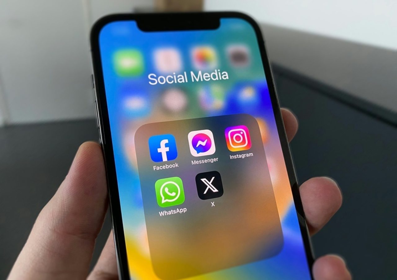 Hand holding a smartphone showing a folder titled Social Media with Facebook, Messenger, Instagram, WhatsApp and X apps visible