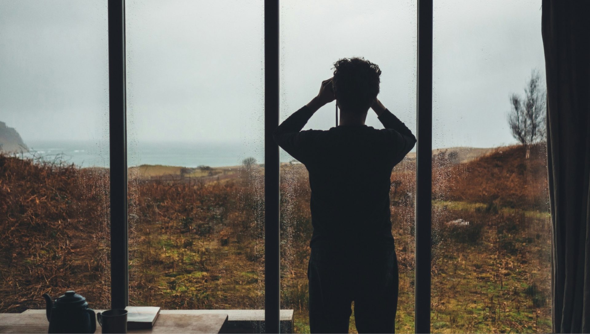 Silhouette of a person looking through a large window with raindrops, overlooking a moody landscape with brown heather and distant sea