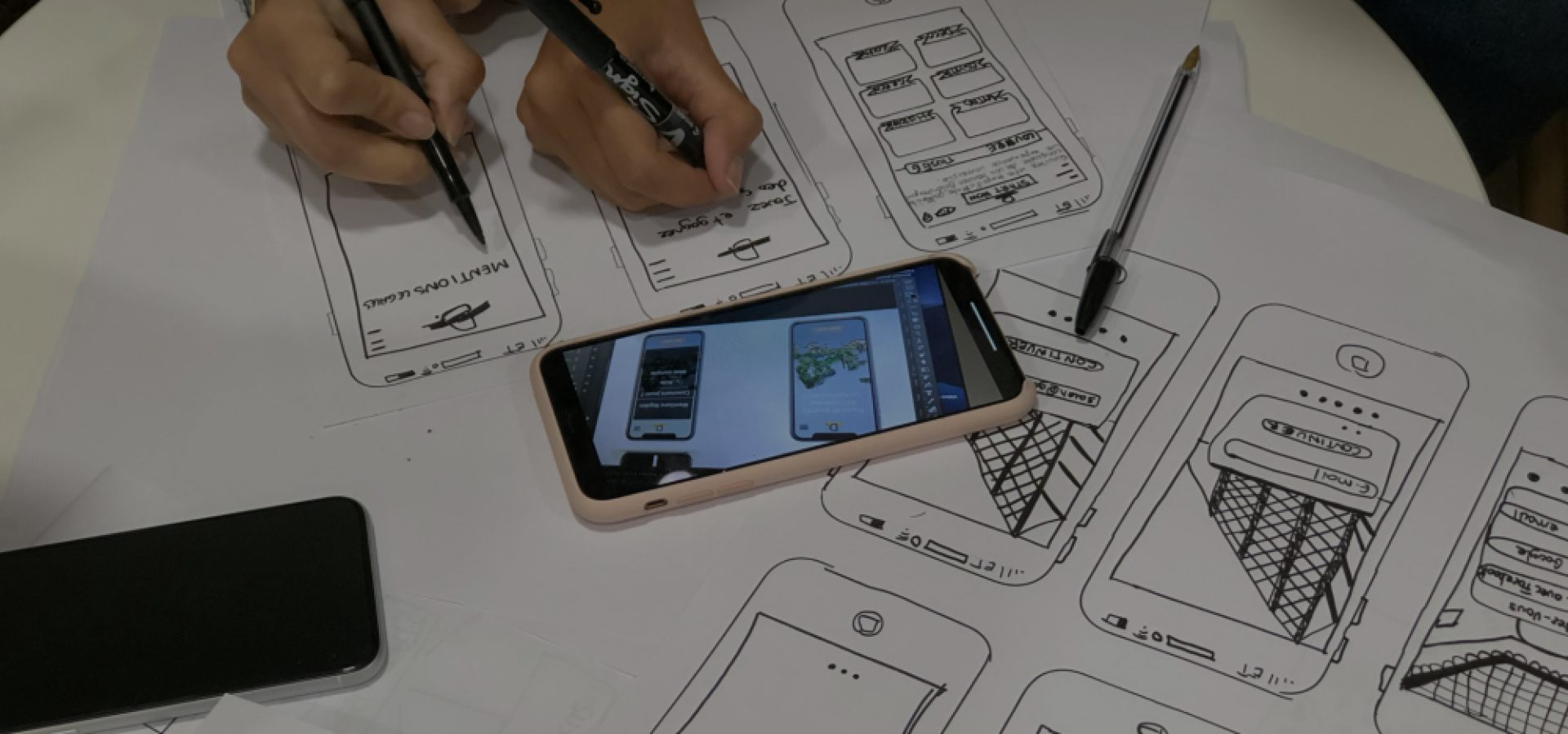 Hands sketching mobile app wireframes on paper with a smartphone displaying digital app designs placed on top