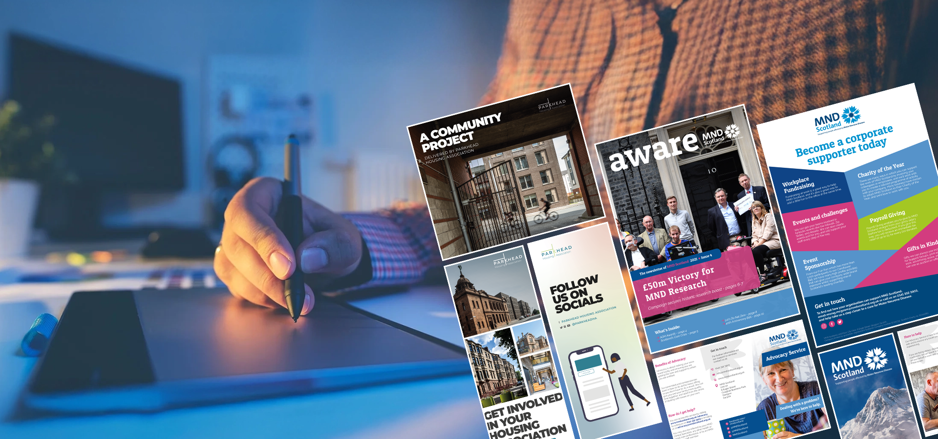 Hand using a digital pen on a tablet with various printed marketing and community project brochures beside it illustrating graphic design work