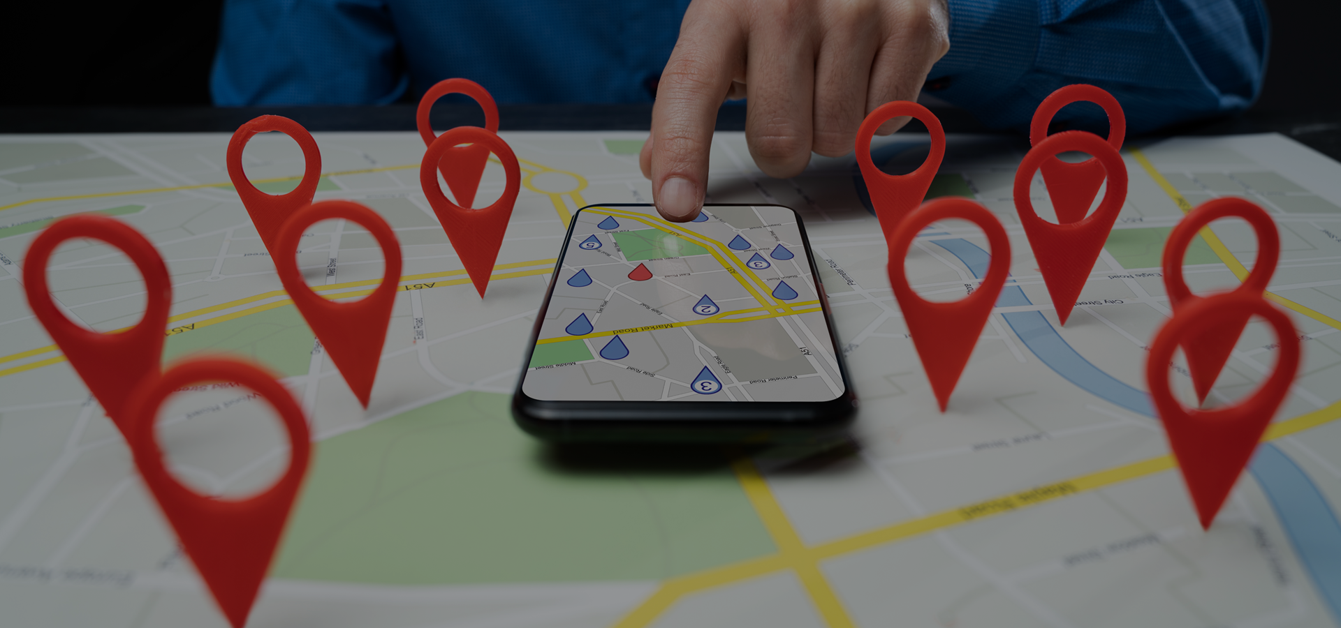 Person using smartphone with map application showing location markers surrounded by large red map pins on a physical map