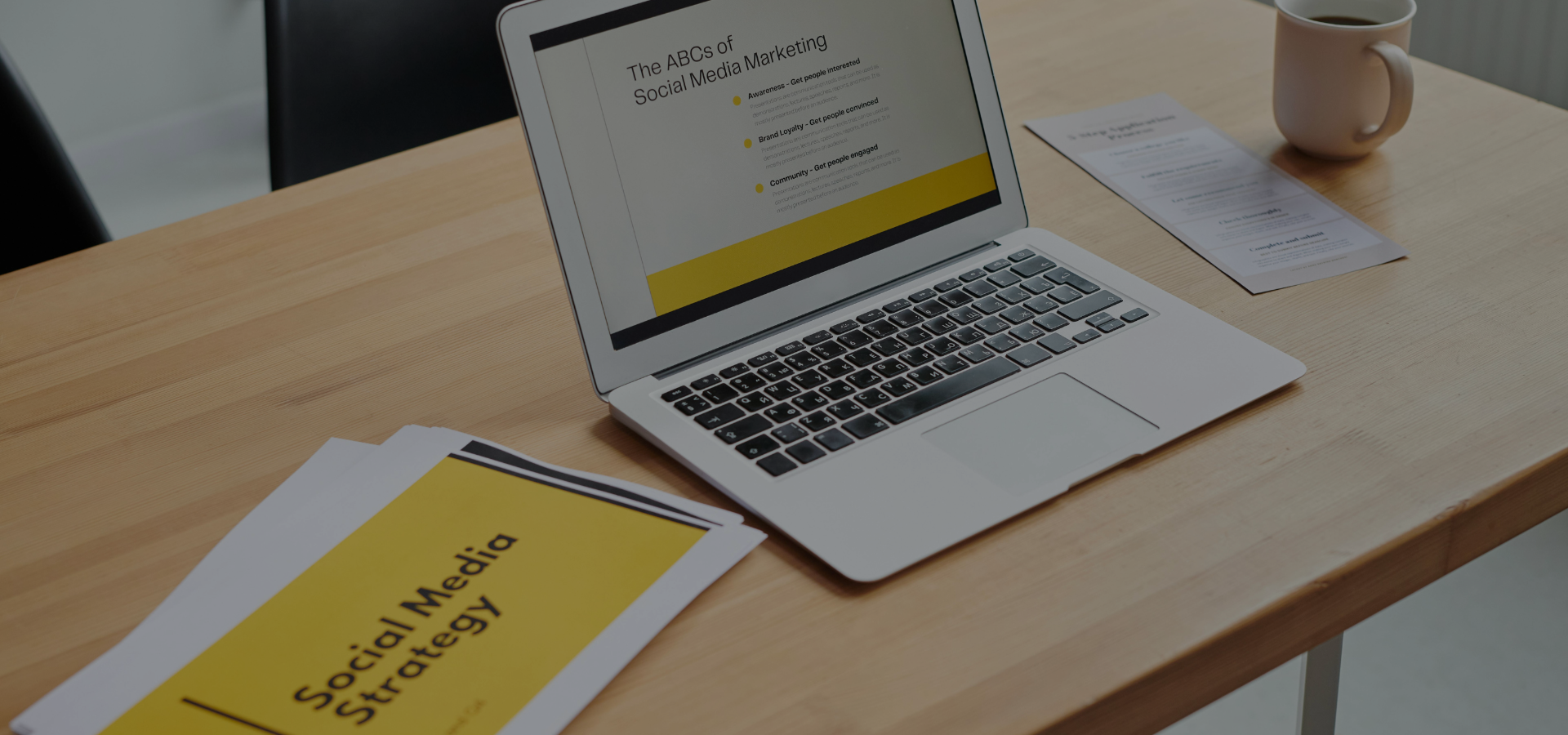 Laptop screen showing a presentation titled 'The ABCs of Social Media Marketing' alongside printed documents titled 'Social Media Strategy' on a wooden desk with a mug