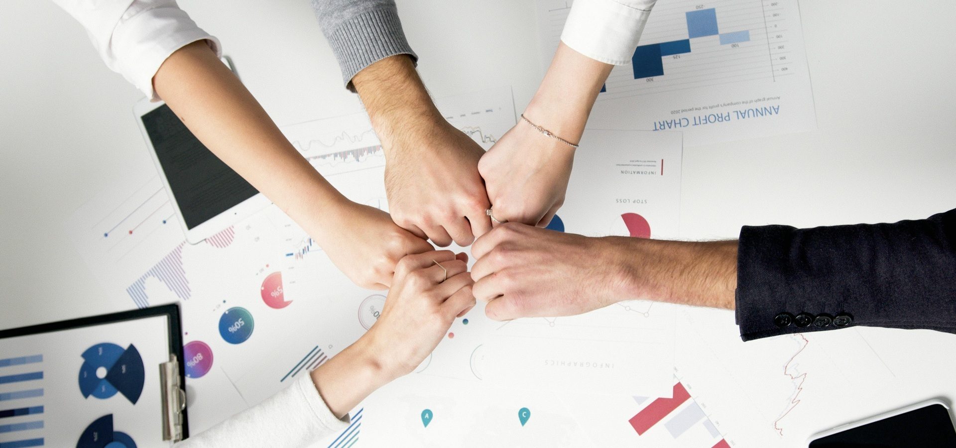 Five people fist bumping over a table covered with printed business charts and graphs, symbolising teamwork and collaboration
