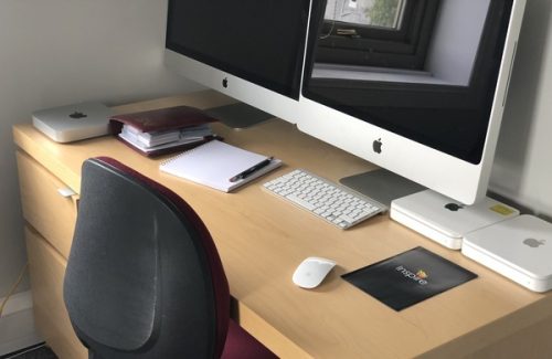Work desk with two Apple iMac computers, wireless keyboard and mouse, notebooks, and Inspire branded mousepad in an office setting