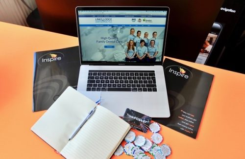 Open laptop on orange desk showing dental practice website surrounded by Inspire-branded brochures, badges, and an open notebook with pen